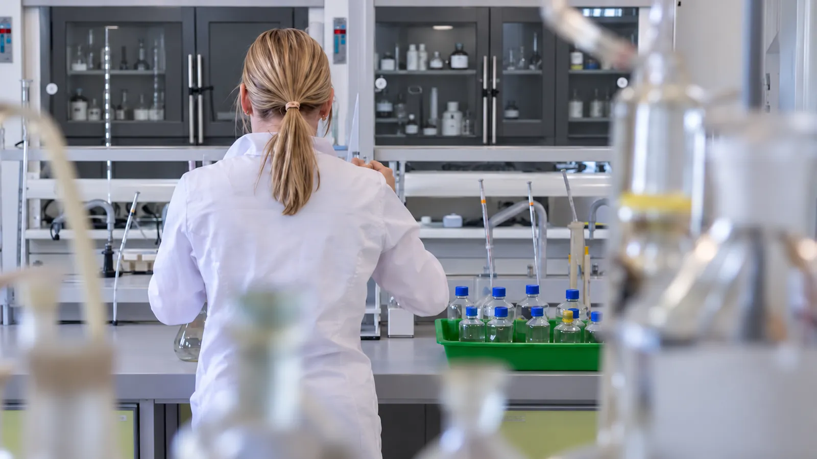 Lab technician in a white coat seen from behind working at a bench beside a green tray of capped glass bottles